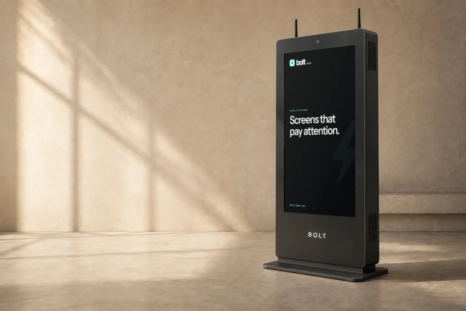 BOLT graphite indoor totem with a vented metal cabinet and a low stepped base in a sunlit stone gallery, showing the “Screens that pay attention.” slide